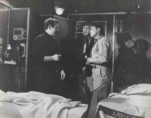 Black-and-white hospital room scene with two beds; a man in dark clothing talks to two men in lighter jackets, as another figure stands in the background.