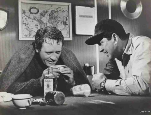Two men sit at a table, one wrapped in a blanket and holding a cup, the other in a cap offers a drink in a dim room with a map on the wall behind them.