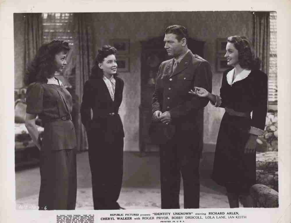 Four adults in vintage attire stand in a living room, with a man in a military uniform center, conversing with three women.