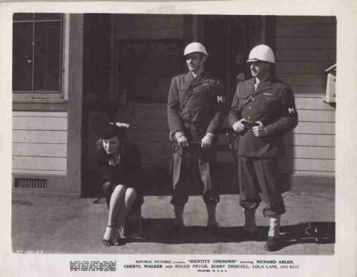 Two World War II-era soldiers in helmets stand with a seated woman in front of a doorway outside a building.