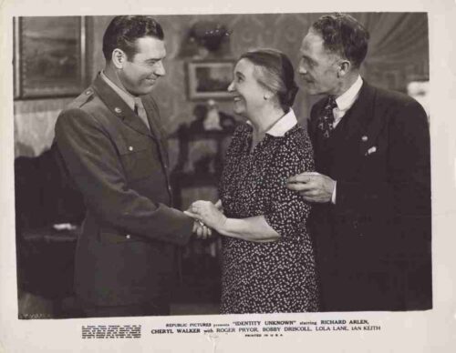 A soldier in uniform shakes hands with a smiling woman while a man in a suit stands behind, holding her arm.
