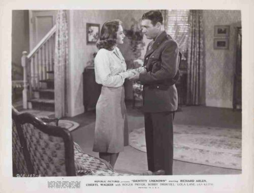 A soldier in uniform holds the woman's hands as they gaze at each other in a living-room setting.