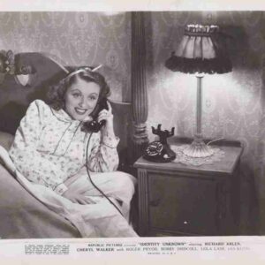 Woman in a floral nightgown sits up in bed, talking on a vintage corded telephone beside a lit bedside lamp and dark wood nightstand to the right.