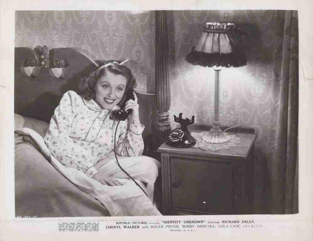 Woman in a floral nightgown sits up in bed, talking on a vintage corded telephone beside a lit bedside lamp and dark wood nightstand to the right.