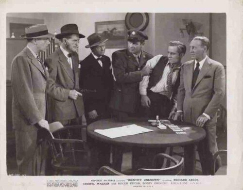 Black-and-white still of six men in suits around a round table; a uniformed officer restrains a man as tension rises around the cards on the table.