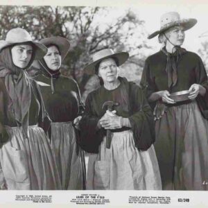Four women in 19th-century dresses and wide-brim hats stand together outdoors in a rural setting, one holding a tool or staff.