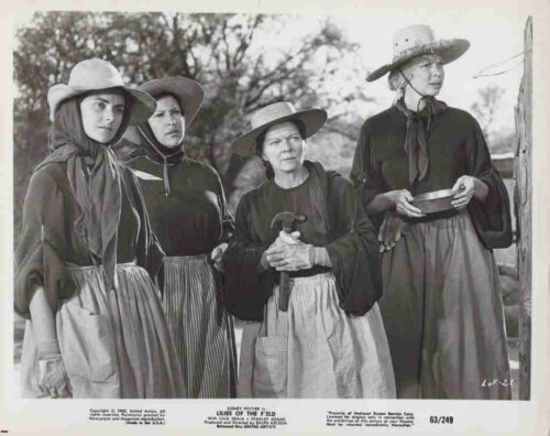 Four women in 19th-century dresses and wide-brim hats stand together outdoors in a rural setting, one holding a tool or staff.