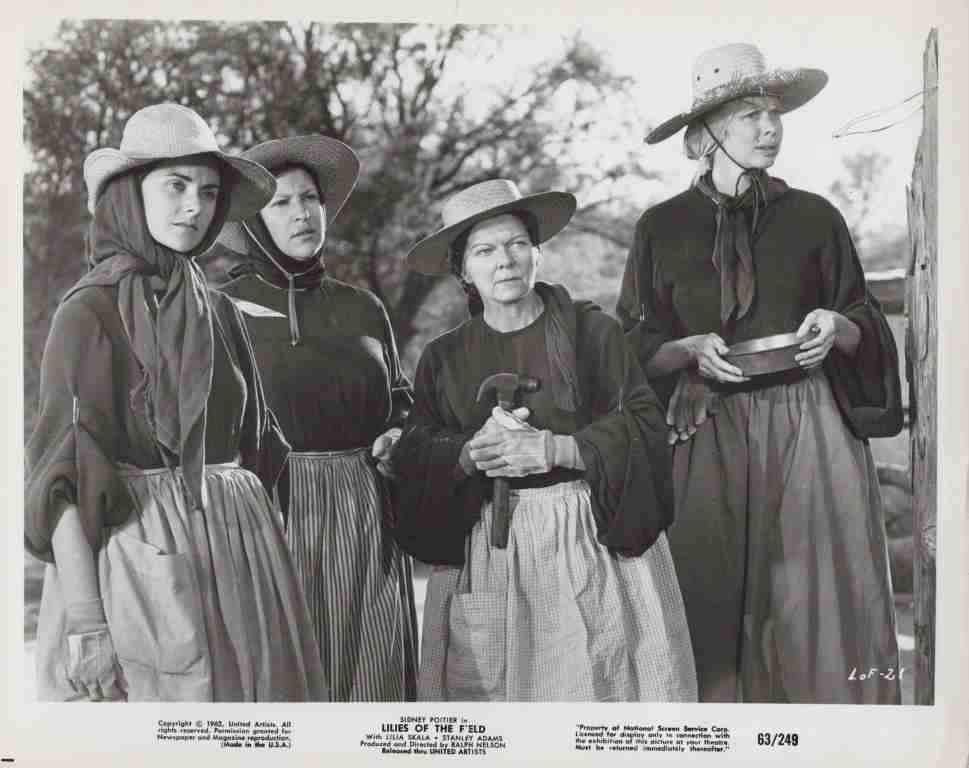 Four women in 19th-century dresses and wide-brim hats stand together outdoors in a rural setting, one holding a tool or staff.