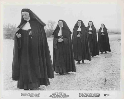 Five nuns in black habits walk single-file along a snowy rural road, heads bowed.