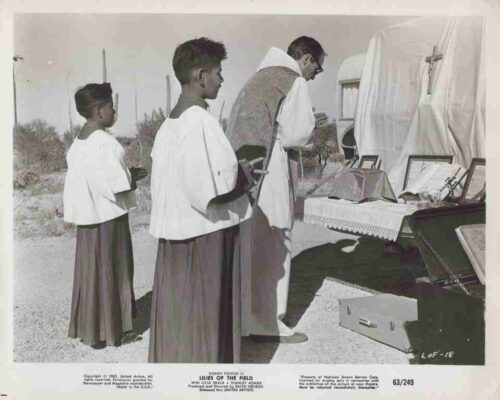 Three men in robes view a table of religious artifacts and framed pictures at an outdoor charity display.] (informative)