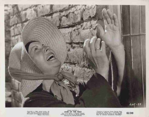 Woman in a large sunhat laughs, leaning against a brick wall with a door, hands raised playfully.