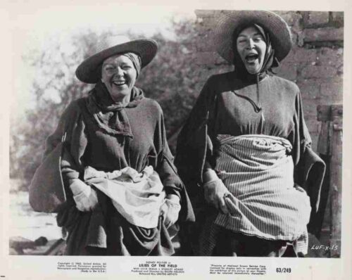 Two elderly women in wide-brim hats laugh together outdoors, wearing traditional plain dresses and aprons against a stone wall backdrop.