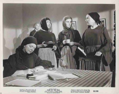 A woman seated at a checkered table signs papers while four other women in headscarves stand nearby, one holding a cup and another with a book, in a room with framed pictures on the wall.