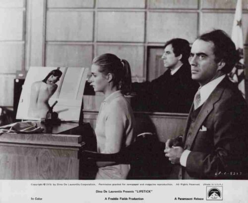 Black-and-white film still: a woman seated at a desk in front of a drawing board with a nude figure sketch, while two men in suits stand behind her in a studio setting.