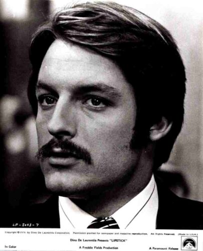 Promotional black-and-white headshot of a man with dark hair and a mustache, wearing a suit and tie, for the film Lipstick.