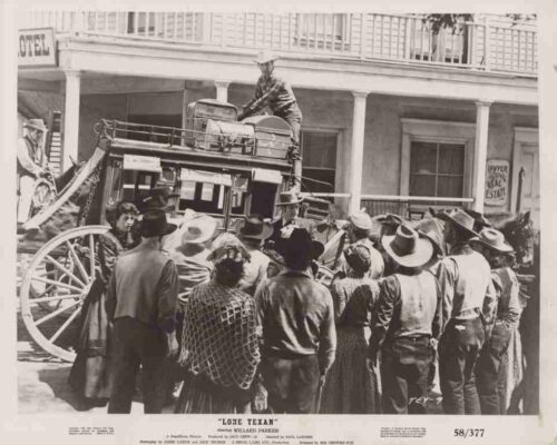 Crowd of 19th-century townspeople surrounds a wheeled stagecoach on a street; a man stands on top of the coach.