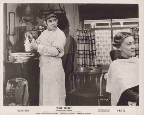 Film still from 'Lone Texan': a man in an apron holding a bottle near a sink, while a woman sits to the right and looks away in a kitchen set.