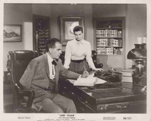 Man seated at an ornate desk signs a document while a woman in a blouse stands beside him in a 1940s-style office with bookshelves in the background .
