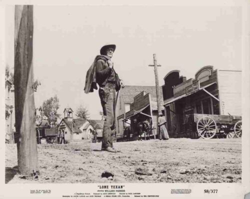 Cowboy in a wide-brimmed hat stands on a dusty Western town street, with wooden storefronts and a wagon in the background.