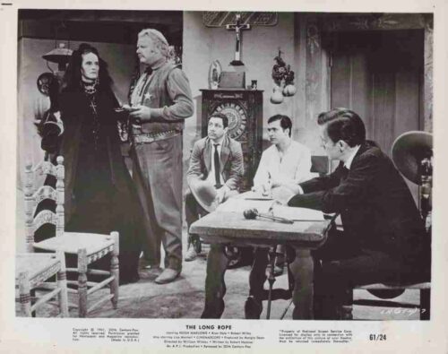 A woman in a dark dress and a bearded man stand in a tense scene while three men sit at a table watching in a cluttered room with a grandfather clock behind them.] - (informative film still)