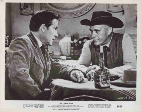 Two men sit at a bar, facing each other and talking, with a bottle on the table between them.