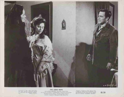 A woman in a light gown stands by a doorway, looking down, as a cloaked figure looms to her left and a man watches from the right side of the room—movie scene.