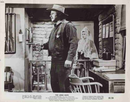 Cowboy in a hat, vest, and checkered shirt aims a pistol in a rustic room; a veiled bride watches from behind him.