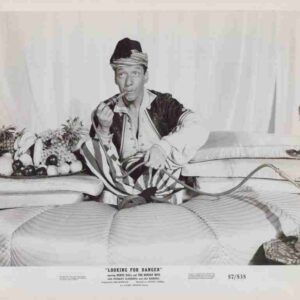 Vintage black-and-white image of a man in a turban lounging on a striped sofa, smoking a pipe, with a fruit platter beside him and a tall hookah on a side table—a posed humorous still from an old stage/promo setting.