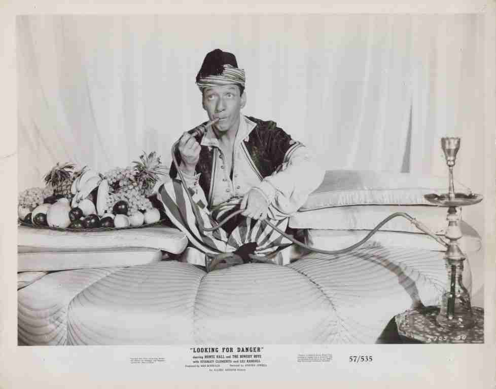 Vintage black-and-white image of a man in a turban lounging on a striped sofa, smoking a pipe, with a fruit platter beside him and a tall hookah on a side table—a posed humorous still from an old stage/promo setting.