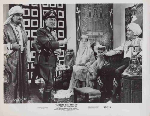 Black-and-white still from a film: a Western officer gestures while a sheikh and a royal entourage sit in an ornately decorated room.