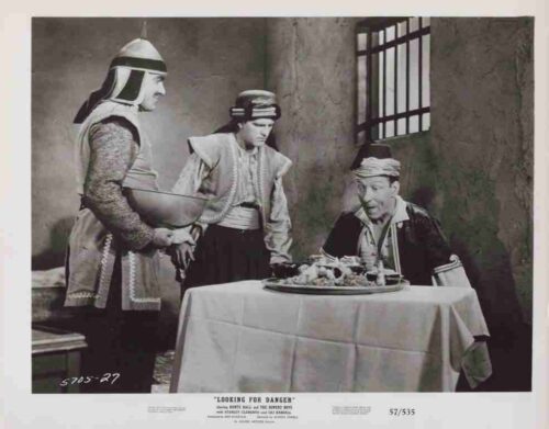 Three men in vintage Middle Eastern costumes stand around a table with a served dish; one man sits, looking intently at the food as the others attend him.