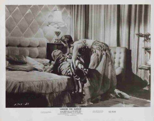 Vintage photo of a man lying on a bed while a woman in an elegant dress leans over him in a luxurious bedroom with a tufted headboard and tall lamp.