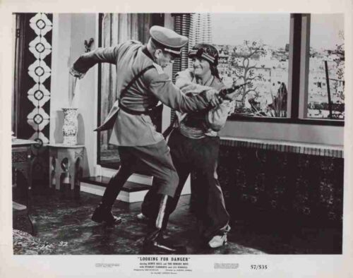 Two men fight in a living room; one in a military-style uniform lunges with a prop pistol toward the other who raises his arms to defend himself in the doorway area of a stylish apartment.