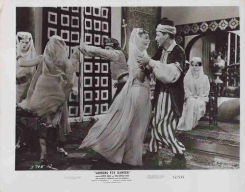 Still from a vintage film scene titled 'Looking for Danger': a man in striped pants dances with a veiled woman while others in veils observe on a richly decorated stage set.
