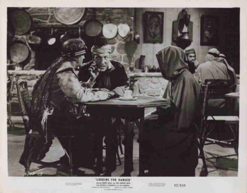 Black-and-white image of a saloon scene: two men at a round table talking, a woman in a hooded cloak nearby, rustic décor and barrels in the background.