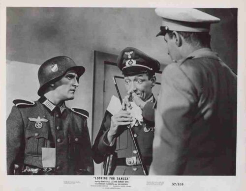 Three men in World War II–era uniforms in a studio: a German officer with a helmet on the left, another German officer in the middle, and a serviceman in a white-caps uniform on the right.