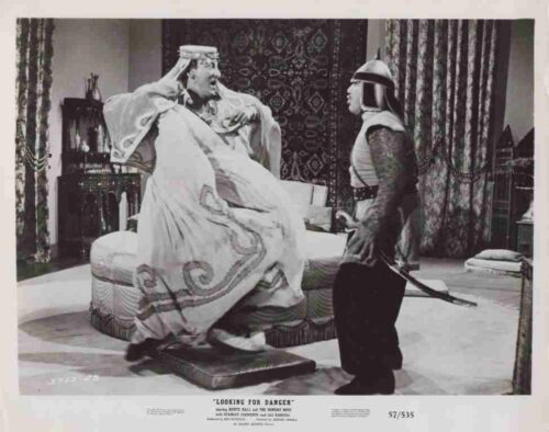 A man in an ornate robe sits up on a throne-like couch as an armored soldier with a helmet advances toward him with a drawn sword in a room with patterned curtains.