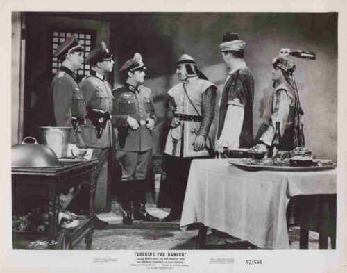 Film still showing six men in period military costumes in a dining room, conversing near a table with food and dishes.
