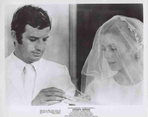 Groom in a white suit places a ring on the bride's finger as she looks down beneath her veil during a wedding.