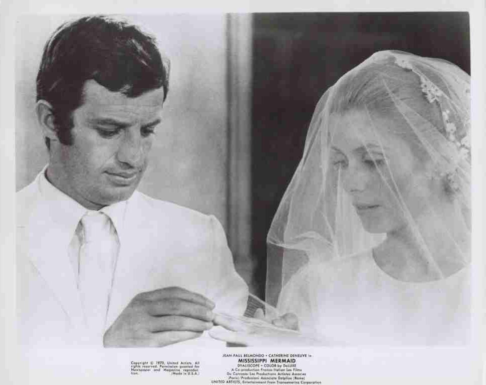 Groom in a white suit places a ring on the bride's finger as she looks down beneath her veil during a wedding.
