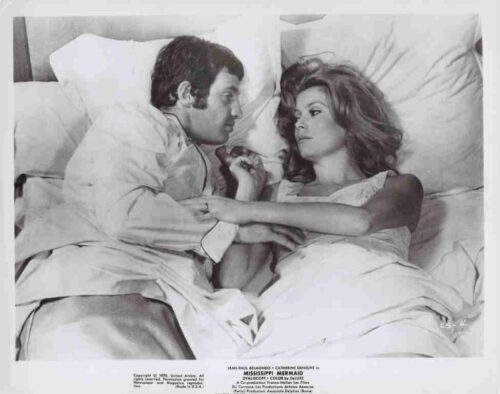 A man and a woman lie in bed facing each other, their hands joined, under white sheets.