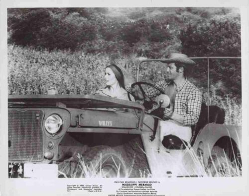 Man wearing a straw hat drives a vintage open-top jeep with a woman passenger in a grassy, rural field. The scene is black-and-white.