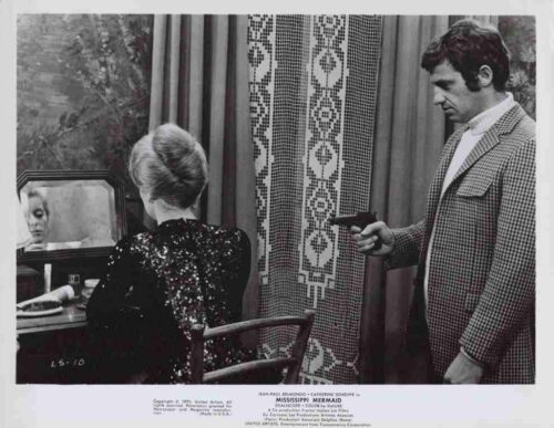Man in a checkered blazer points a pistol at a seated woman in a sequined dress, in a dressing-room style setting with a mirror nearby.