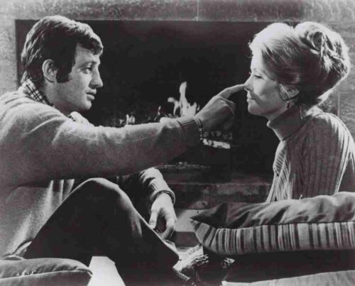 Man sits on a couch by a lit fireplace, touching a woman’s nose playfully.