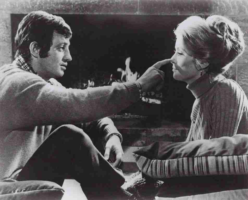 Man sits on a couch by a lit fireplace, touching a woman’s nose playfully.