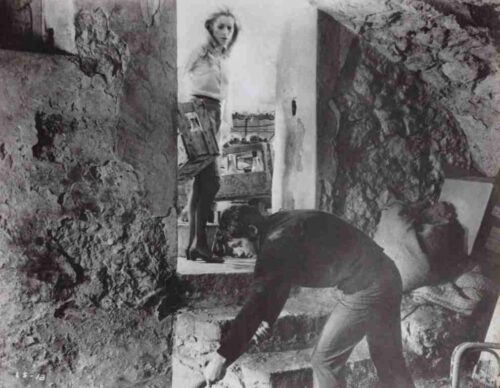 Two people inside a rough, cave-like room; one crouches to work on the stone floor while the other stands in a doorway.