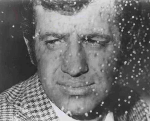Close-up of a man’s face with a serious expression, seen through rain-speckled glass on a black-and-white photo.