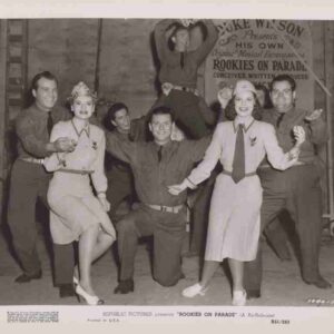 Group of performers in vintage military-style costumes posing on a film set; two women in light dresses stand front and center while others strike playful poses around them, a banner behind reads 'Rookies on Parade'.