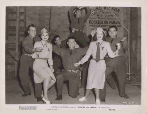 Group of performers in vintage military-style costumes posing on a film set; two women in light dresses stand front and center while others strike playful poses around them, a banner behind reads 'Rookies on Parade'.