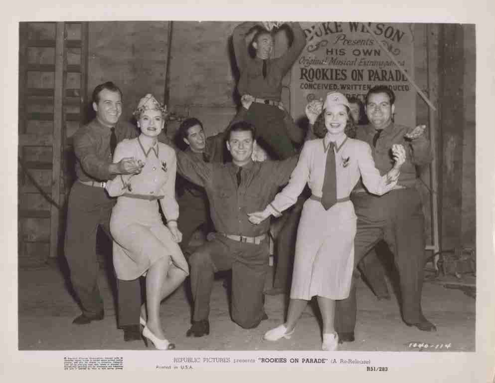 Group of performers in vintage military-style costumes posing on a film set; two women in light dresses stand front and center while others strike playful poses around them, a banner behind reads 'Rookies on Parade'.